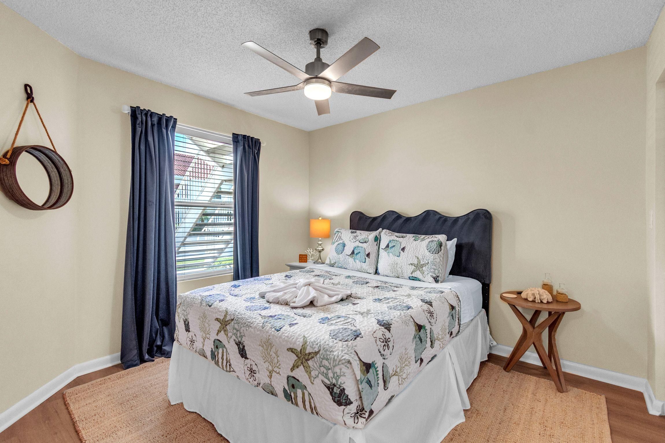 A bedroom with a bed, a ceiling fan, a window with curtains, a bedside lamp, and a small wooden table.
