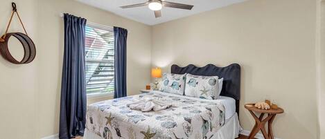Un dormitorio con cama, ventilador de techo, ventana con cortinas, lámpara de noche y una pequeña mesa de madera.