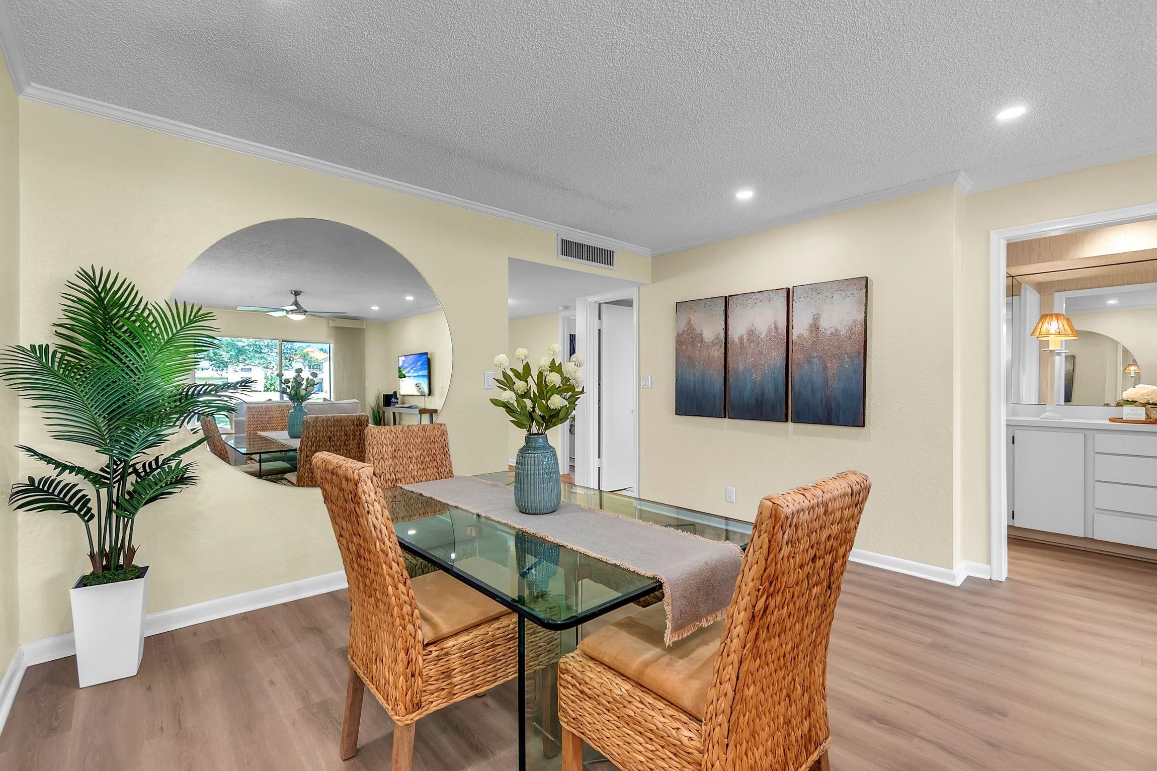 A dining area with a glass table, woven chairs, a vase with flowers, and a view of the living room.