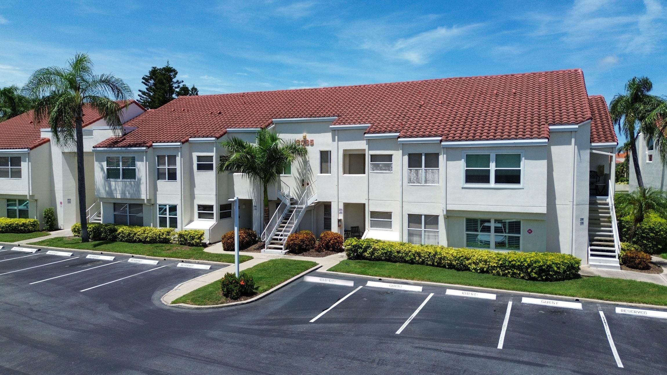 A multi-story residential building with a red-tiled roof, a parking lot with marked guest and reserved spaces, and a staircase leading to the entrance.