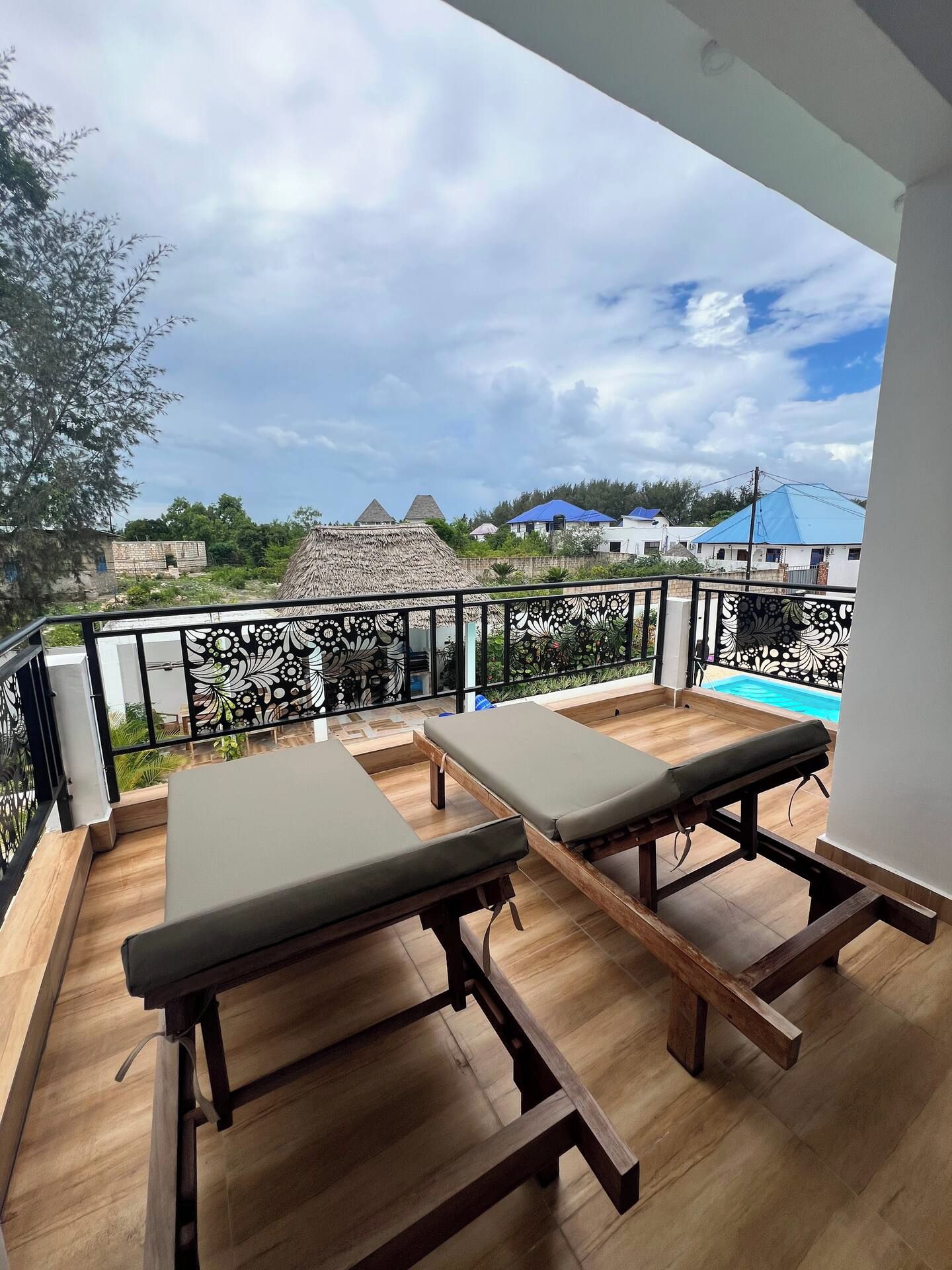 Uma varanda com duas poltronas de descanso, piscina e vista para uma aldeia com casas e uma construção com telhado de palha.