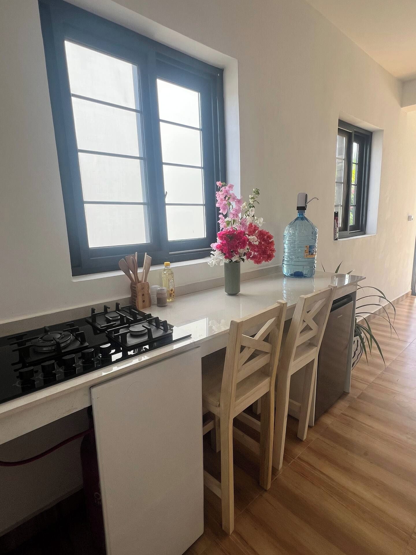 A kitchen with a gas stove, wooden chairs, a large water dispenser, and a window with a view.