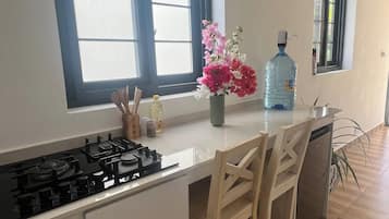 A kitchen with a gas stove, wooden chairs, a large water dispenser, and a window with a view.