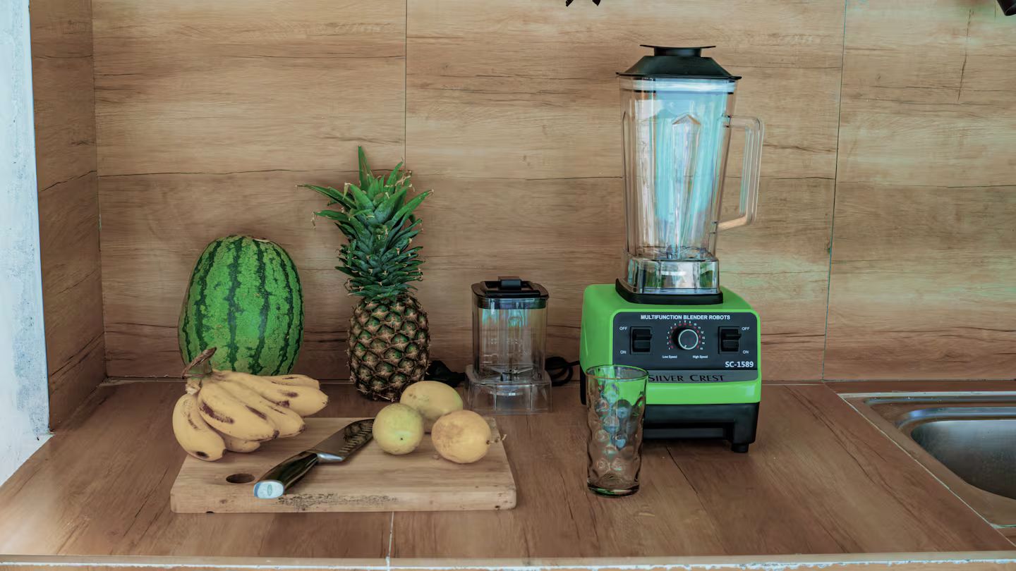 A kitchen counter with a blender, a cutting board with fruits, and a glass.