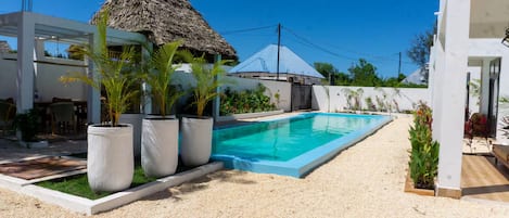 A swimming pool with a white deck, surrounded by potted plants and a thatched-roof structure.