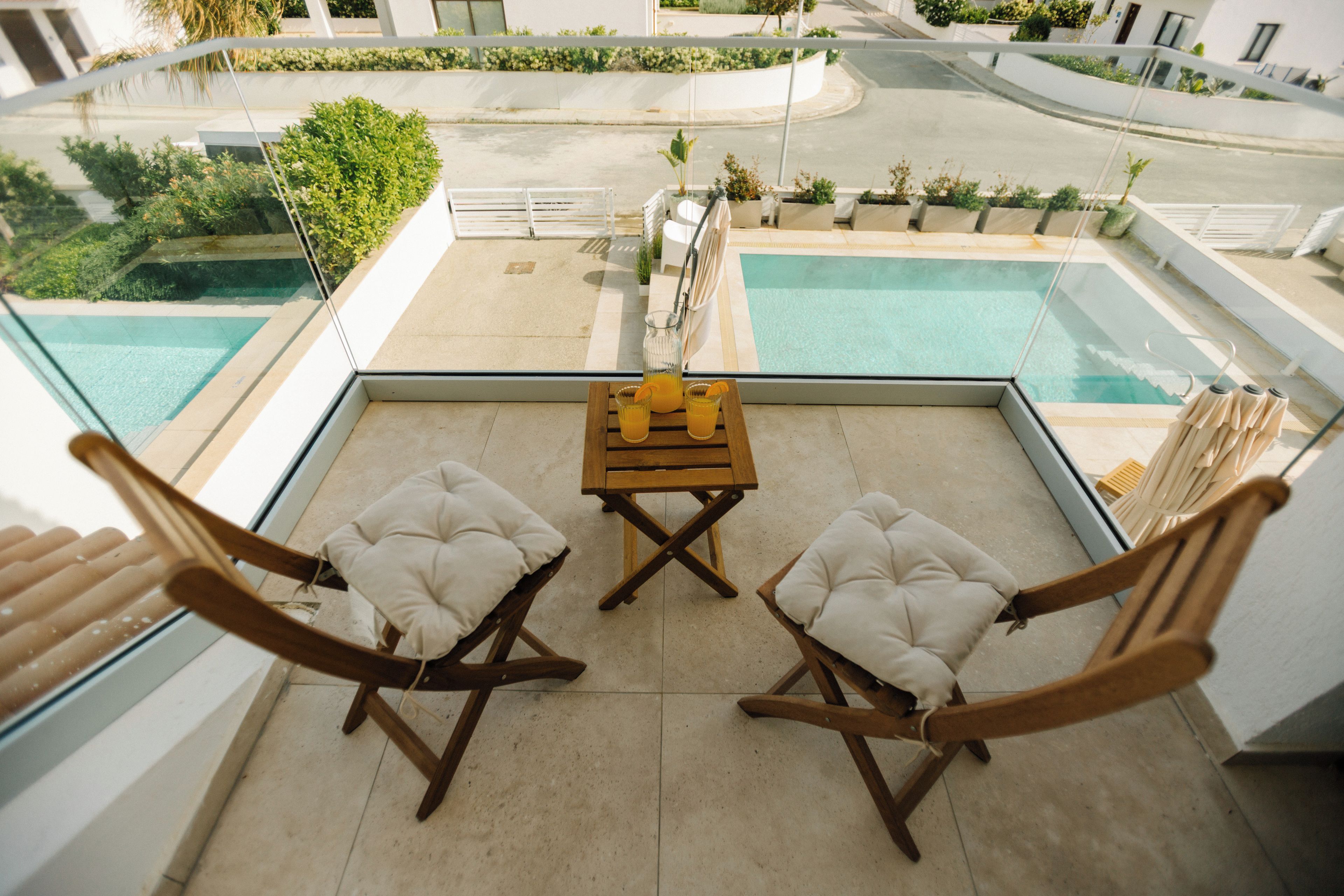 A poolside area with two wooden chairs and a table, overlooking a swimming pool and a landscaped garden.