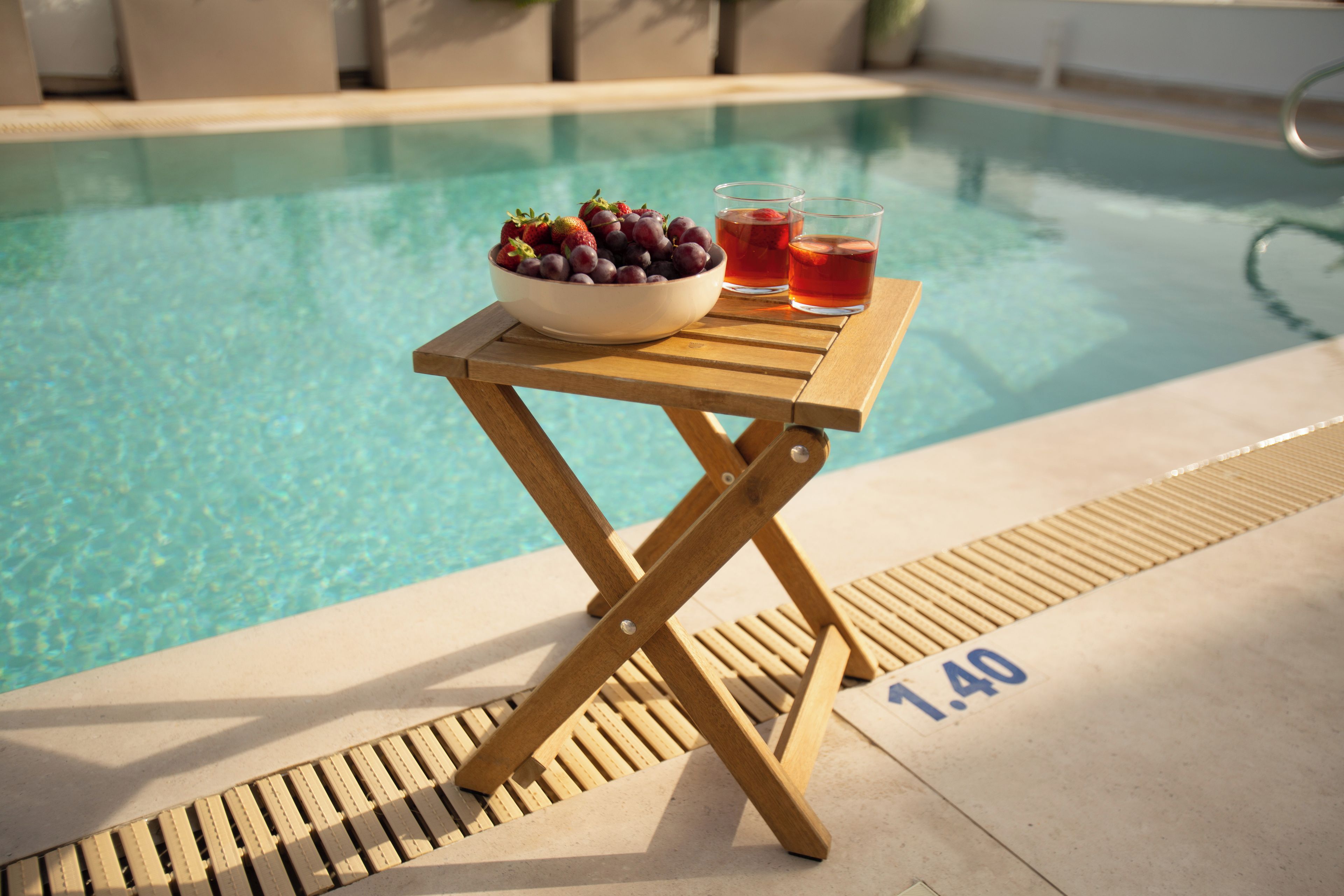 A poolside table with a bowl of fruit and drinks, a pool in the background, and a lifeguard sign.