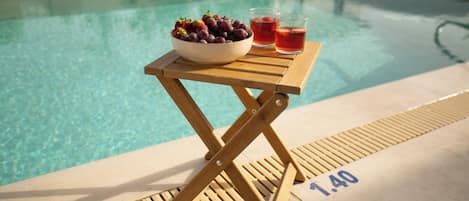 A poolside table with a bowl of fruit and drinks, a pool in the background, and a lifeguard sign.
