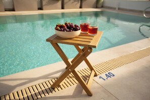 Et bord ved poolen med en skål frugt og drikkevarer, en pool i baggrunden og et skilt med livredder.