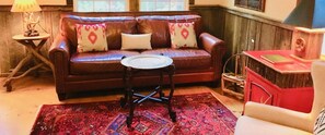 A brown leather sofa with patterned cushions, a small round table, a red patterned rug, and a wooden birdhouse on a red cabinet. - Redesigned Mountain Cottage w/ Fireplace & Barn Wood Accents — Sleeps 4 (Lakemont)