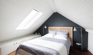 Ein Schlafzimmer mit schrägem Dach, einem Bett mit Holz-Kopfteil, Nachttischlampen und einem Oberlicht.