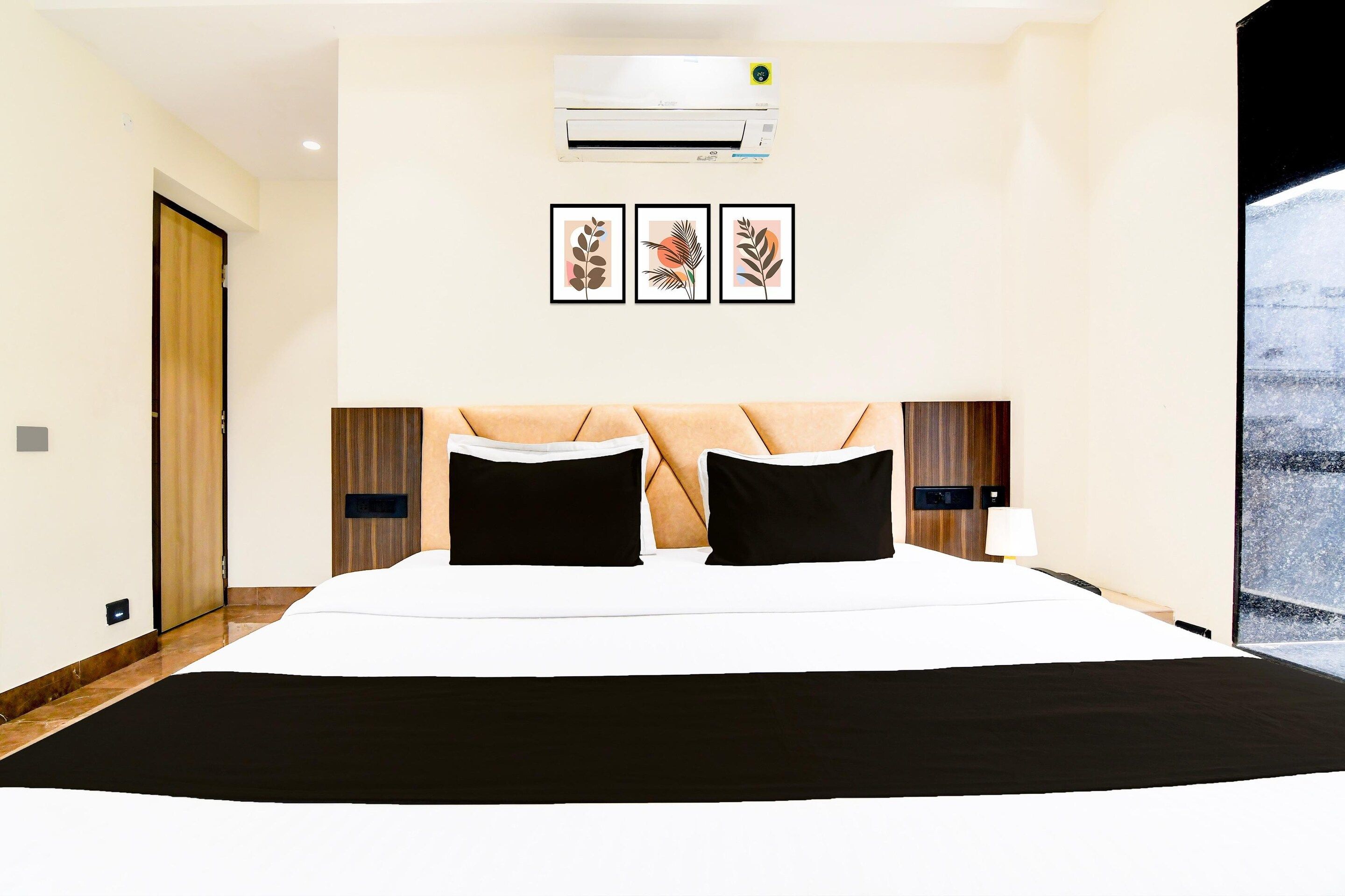 A hotel room with a large bed, an air conditioner, framed artwork on the wall, and a window with a view.