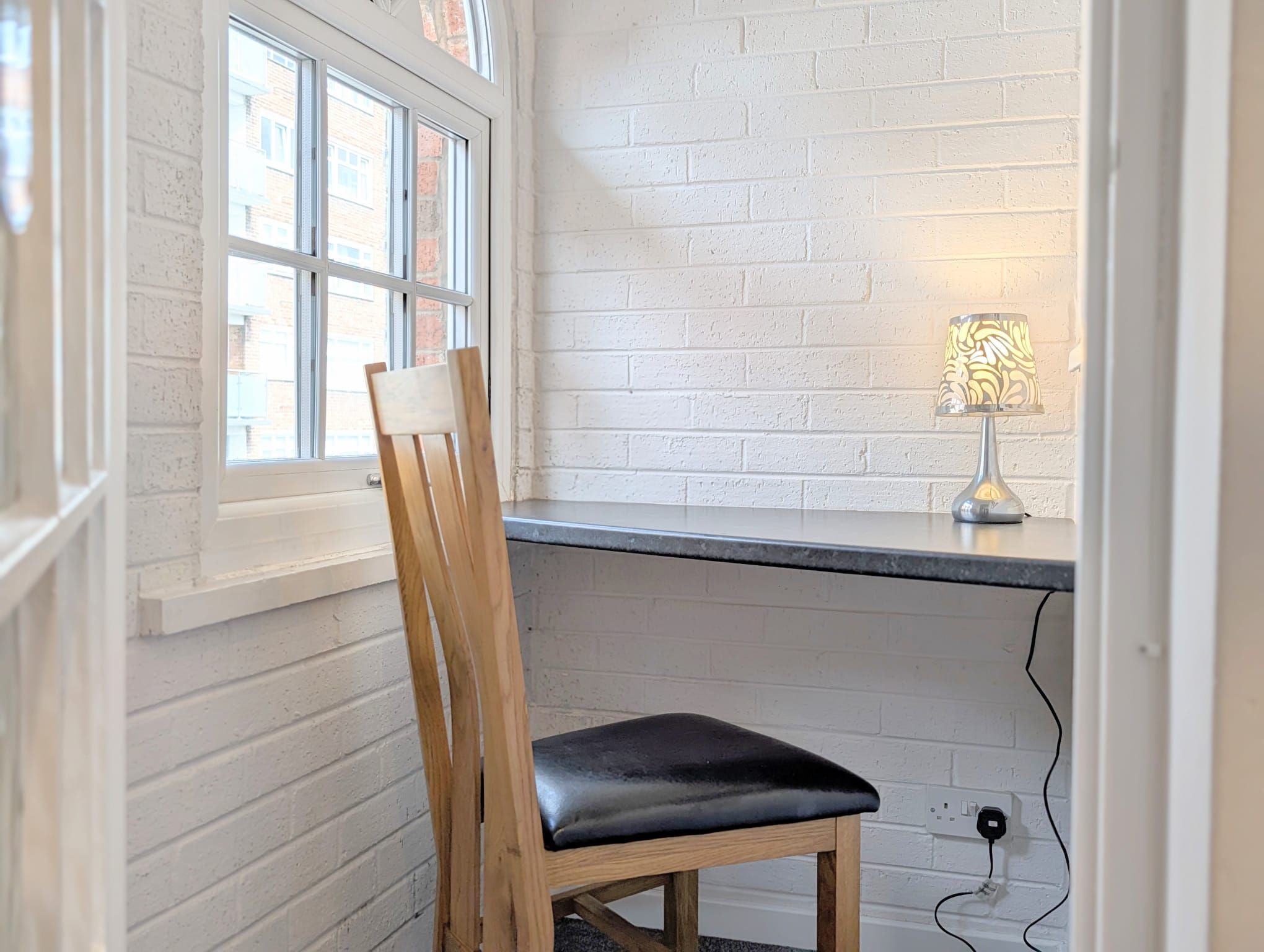 A corner room with a white brick wall, a window, a desk with a lamp, and a wooden chair.