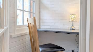 A corner room with a white brick wall, a window, a desk with a lamp, and a wooden chair.