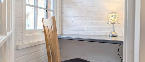 A corner room with a white brick wall, a window, a desk with a lamp, and a wooden chair.