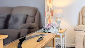 A living room with a sofa, a TV on a stand, a remote control, a lamp, and a small table with flowers. - Stylish Southport Town Centre Retreat + Parking (Southport)