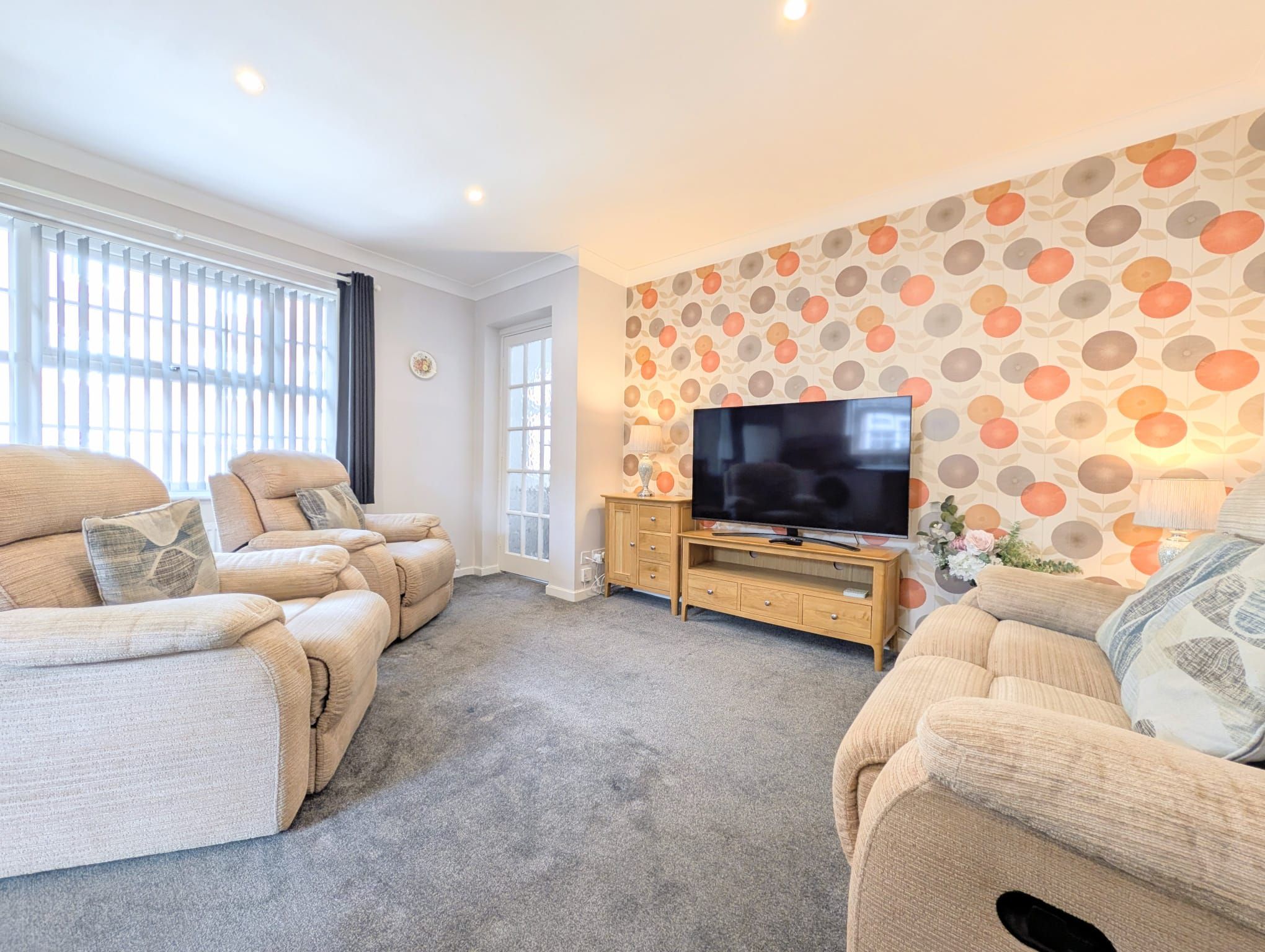 A living room with a sofa set, a TV on a wooden stand, and a patterned wall.