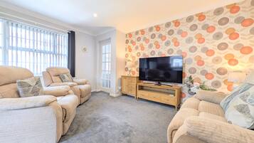 A living room with a sofa set, a TV on a wooden stand, and a patterned wall.