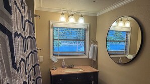 Un baño con tocador, espejo, ventana con persianas, toallero y dispensador de papel higiénico.