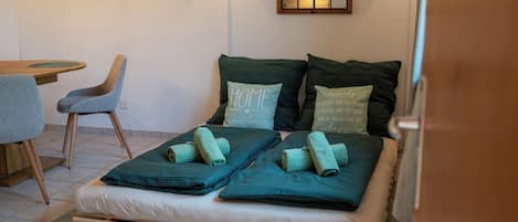 Un lit simple avec deux oreillers, une tête de lit en bois et une petite table avec une chaise, dans une pièce aux murs blancs et dotée d’une fenêtre.