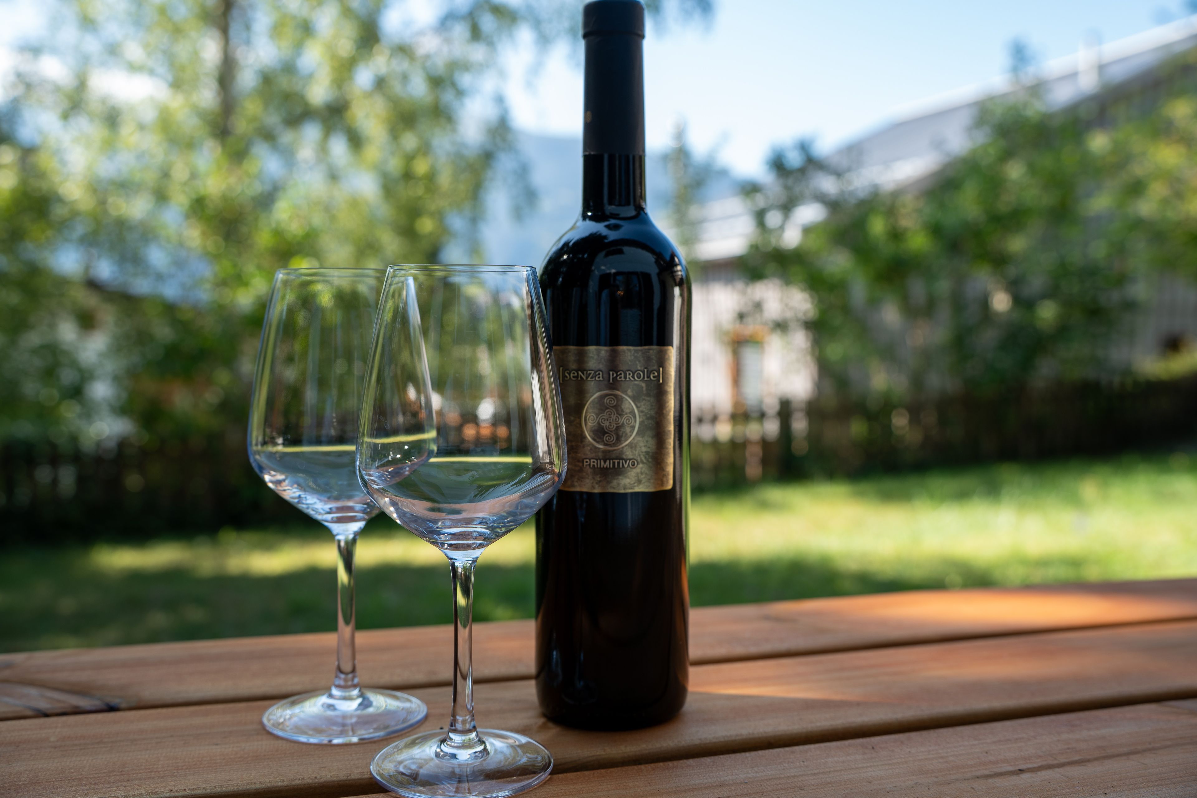 A bottle of wine and two glasses on a wooden table outdoors.