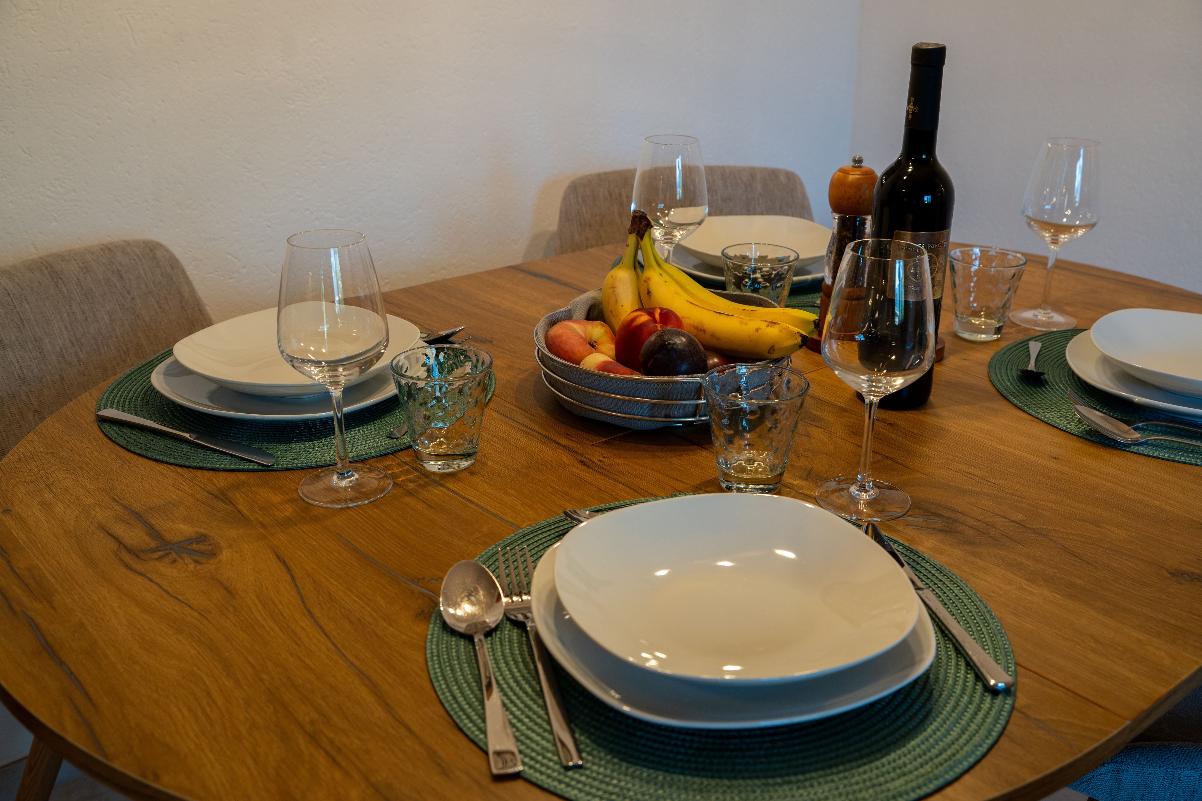 Mesa posta com pratos, copos, talheres, uma garrafa e uma tigela de frutas.