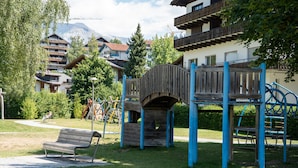 En lekplats med en träbro, en bänk och utsikt över bostadshus och berg i bakgrunden.