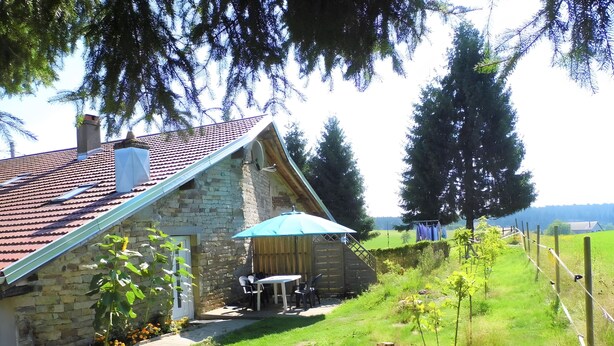 A stone house with a tiled roof, a wooden deck, and a small table with chairs. - Maison Principale Apartment: Mountain View, Private Terrace, and Wi-Fi (Xertigny)