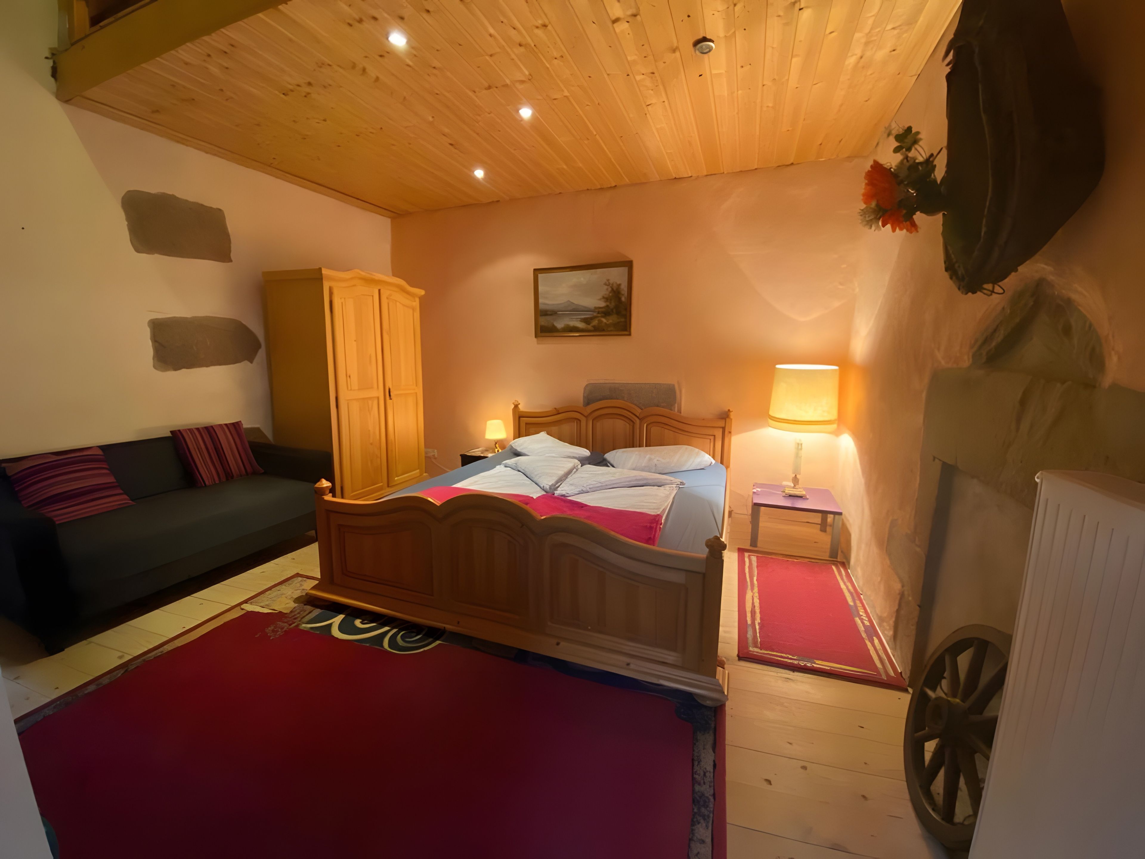 Een slaapkamer met een houten plafond, een bed met een rood-blauwe dekbedovertrek, een nachtkastje met een lamp, een houten kledingkast, en een radiator aan de muur.
