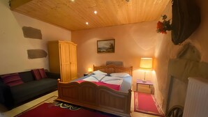 A bedroom with a wooden ceiling, a bed with a red and blue bedspread, a nightstand with a lamp, a wooden wardrobe, and a wall-mounted radiator. - Maison Principale Apartment: Mountain View, Private Terrace, and Wi-Fi (Xertigny)