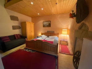 Een slaapkamer met een houten plafond, een bed met een rood-blauwe dekbedovertrek, een nachtkastje met een lamp, een houten kledingkast, en een radiator aan de muur.