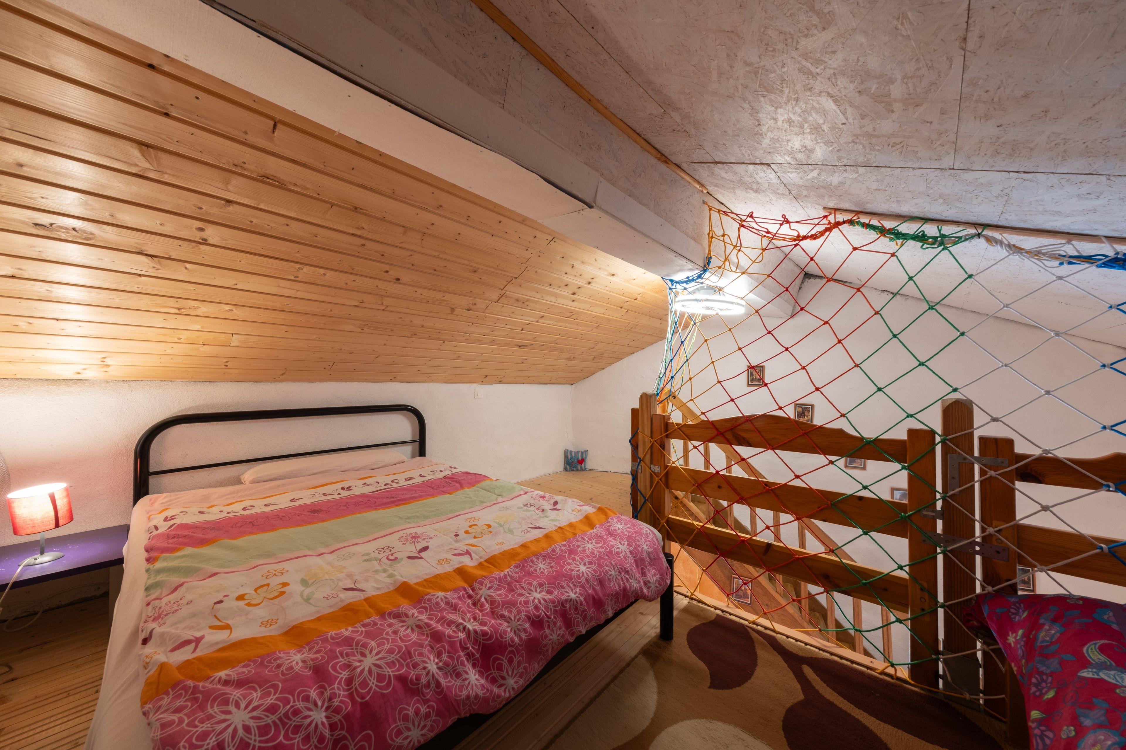 A bedroom with a wooden ceiling, a bed with a colorful quilt, a bedside table with a lamp, and a window with a view of the outdoors.