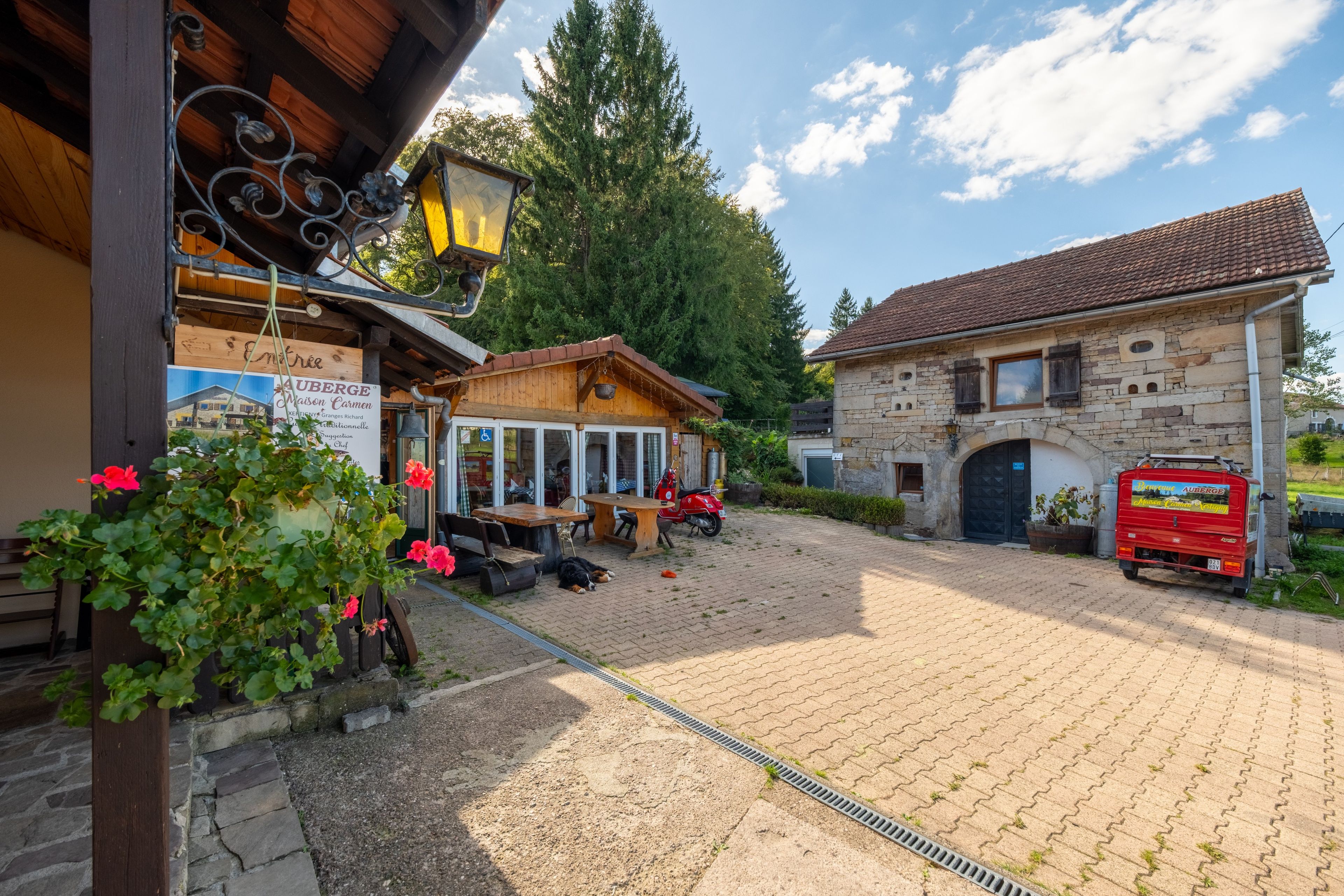 A courtyard with a wooden building, a stone building, and a red vehicle parked outside.