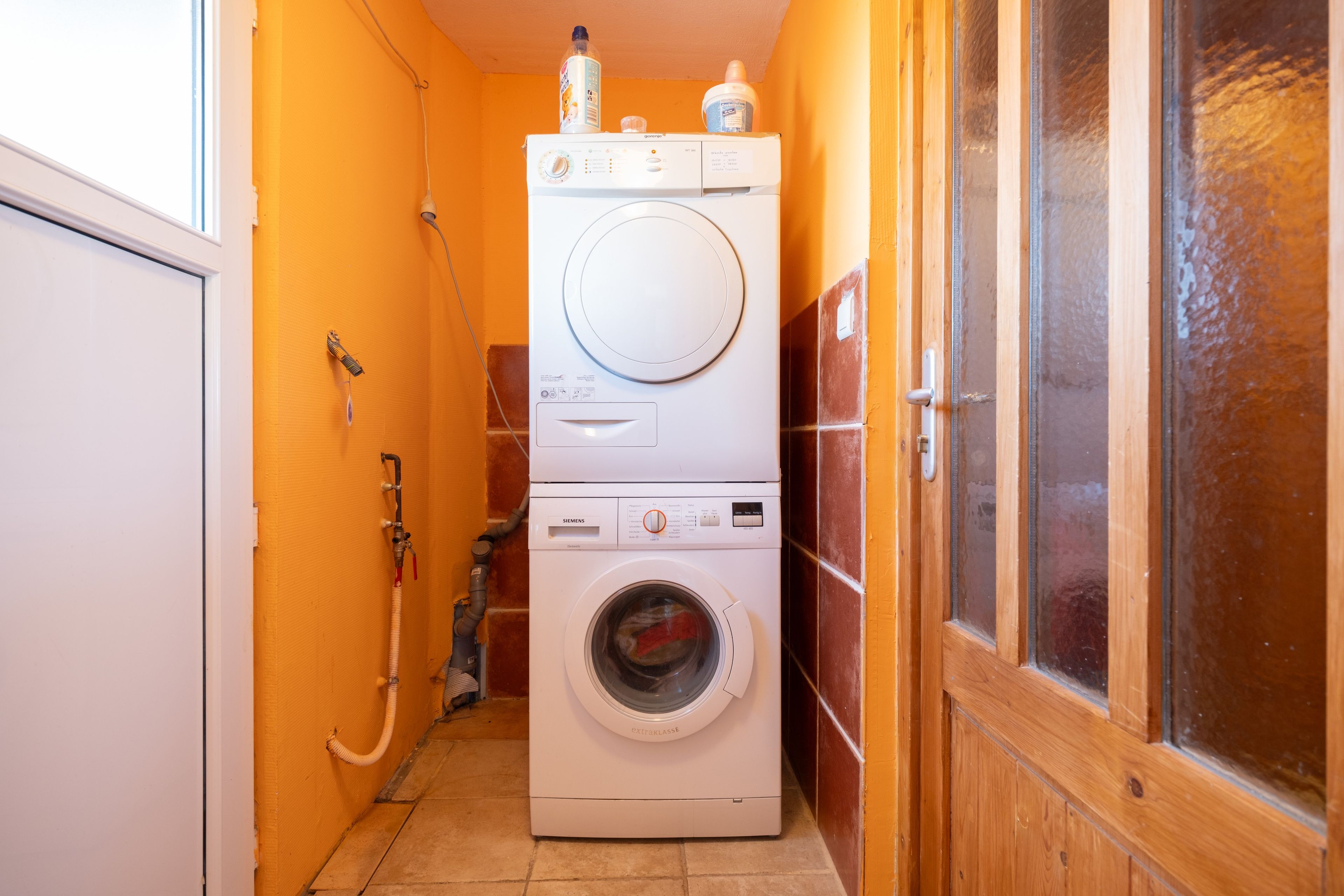 Uma lavanderia compacta com máquina de lavar e secadora empilhadas, uma mangueira e uma porta de madeira.