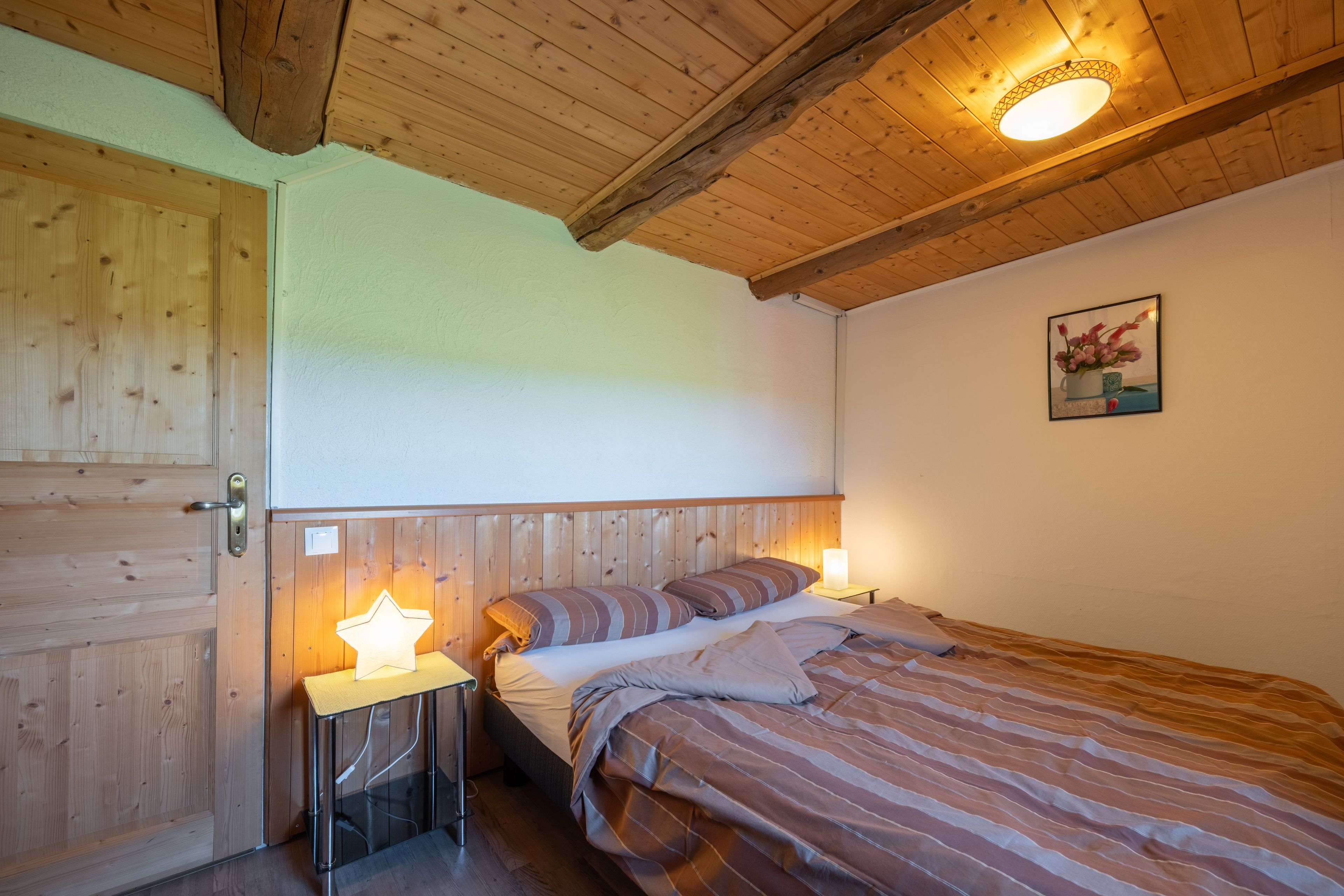 Quarto com teto de madeira, cama com roupa de cama listrada, mesa de cabeceira com abajur em formato de estrela e um quadro na parede.