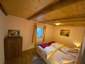 Ein Schlafzimmer mit Holzboden, einem fenster mit Vorhängen, einem Bett mit vielen Kissen, einem Nachttisch mit Lampe und einem Schrank mit einem Gemälde.