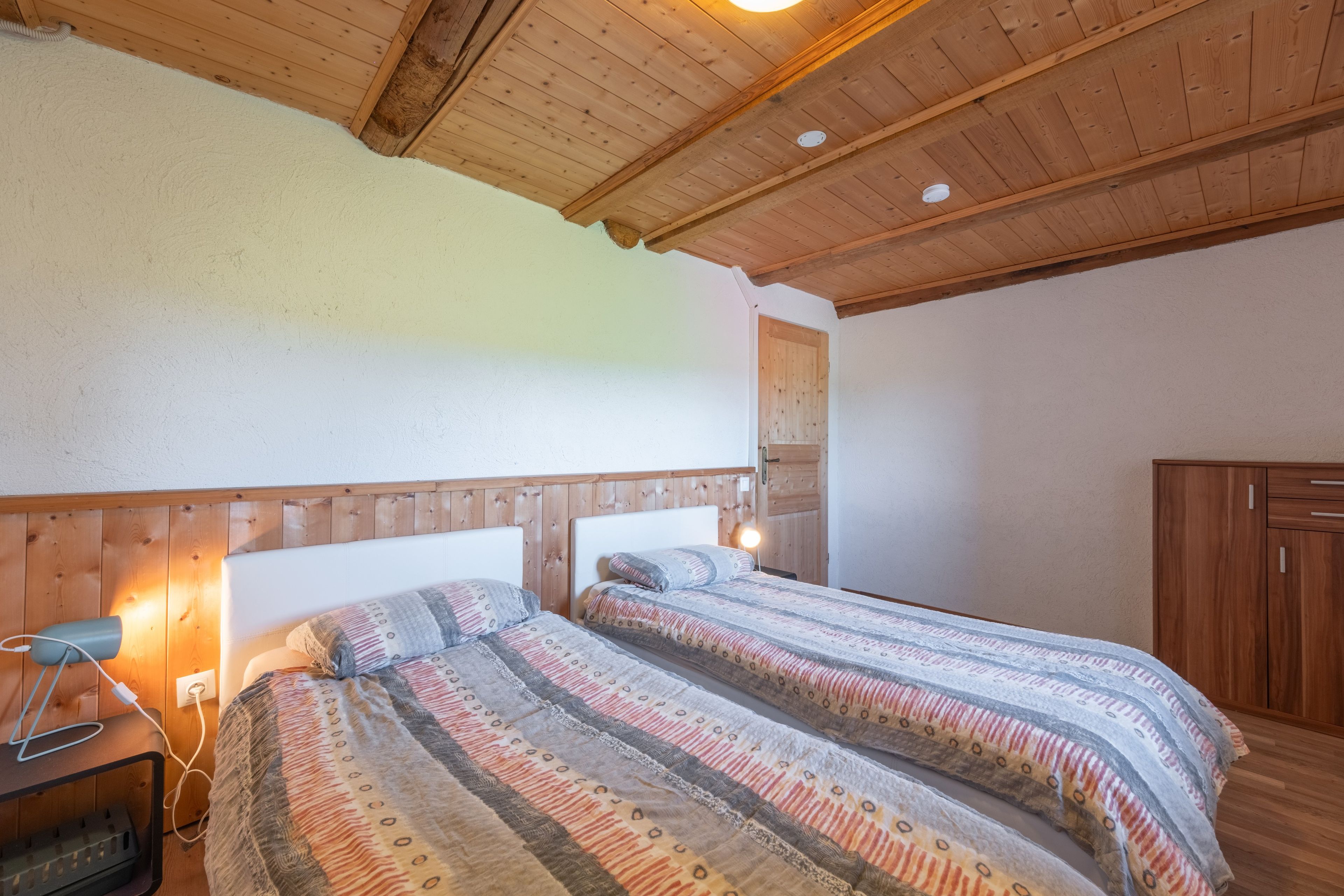 Un dormitorio con dos camas, cabeceros de madera, una mesita de noche con lámpara y un armario.
