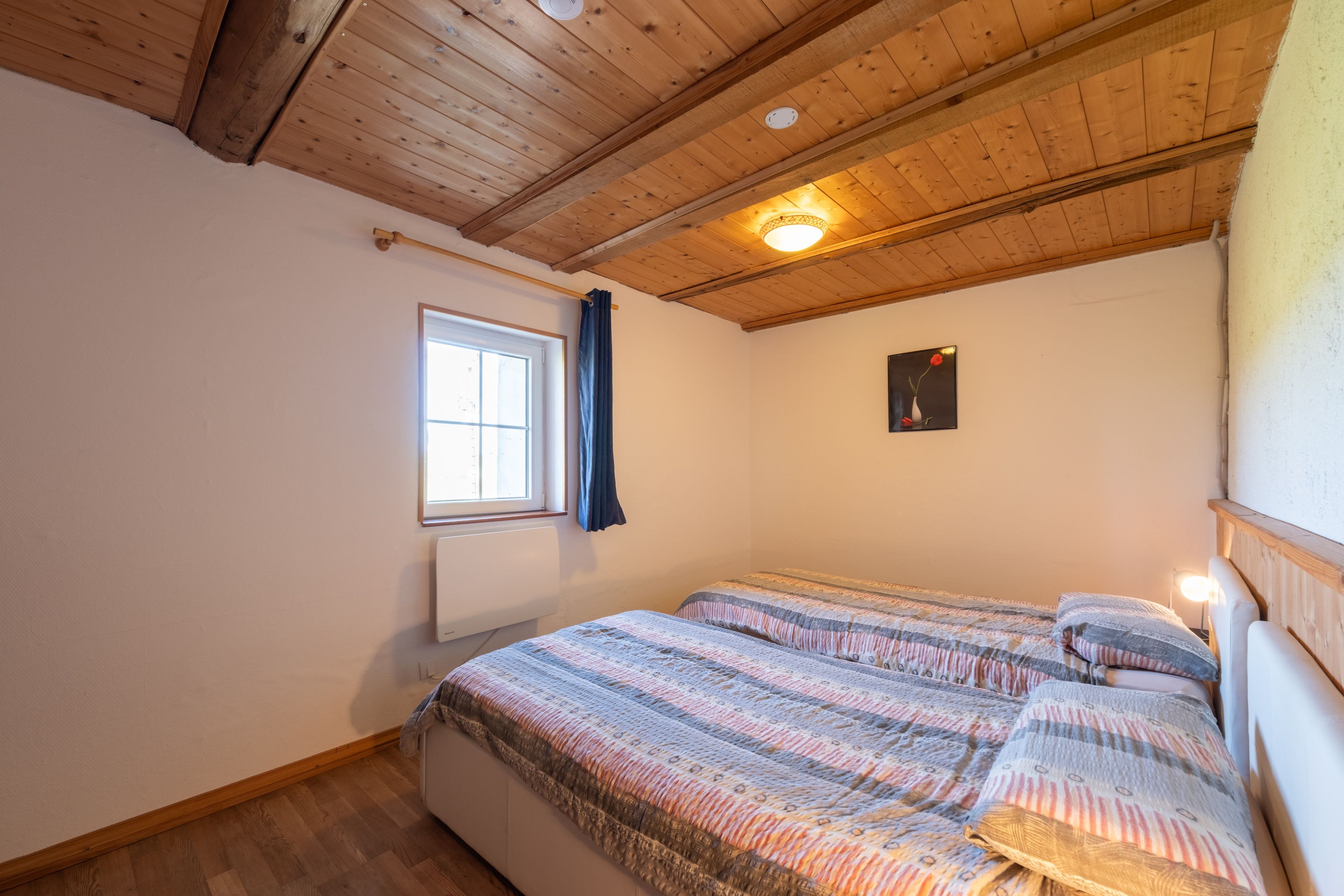 Une pièce avec un plafond en bois, une fenêtre avec des rideaux, un lit avec une literie à motifs et un tableau accroché au mur.