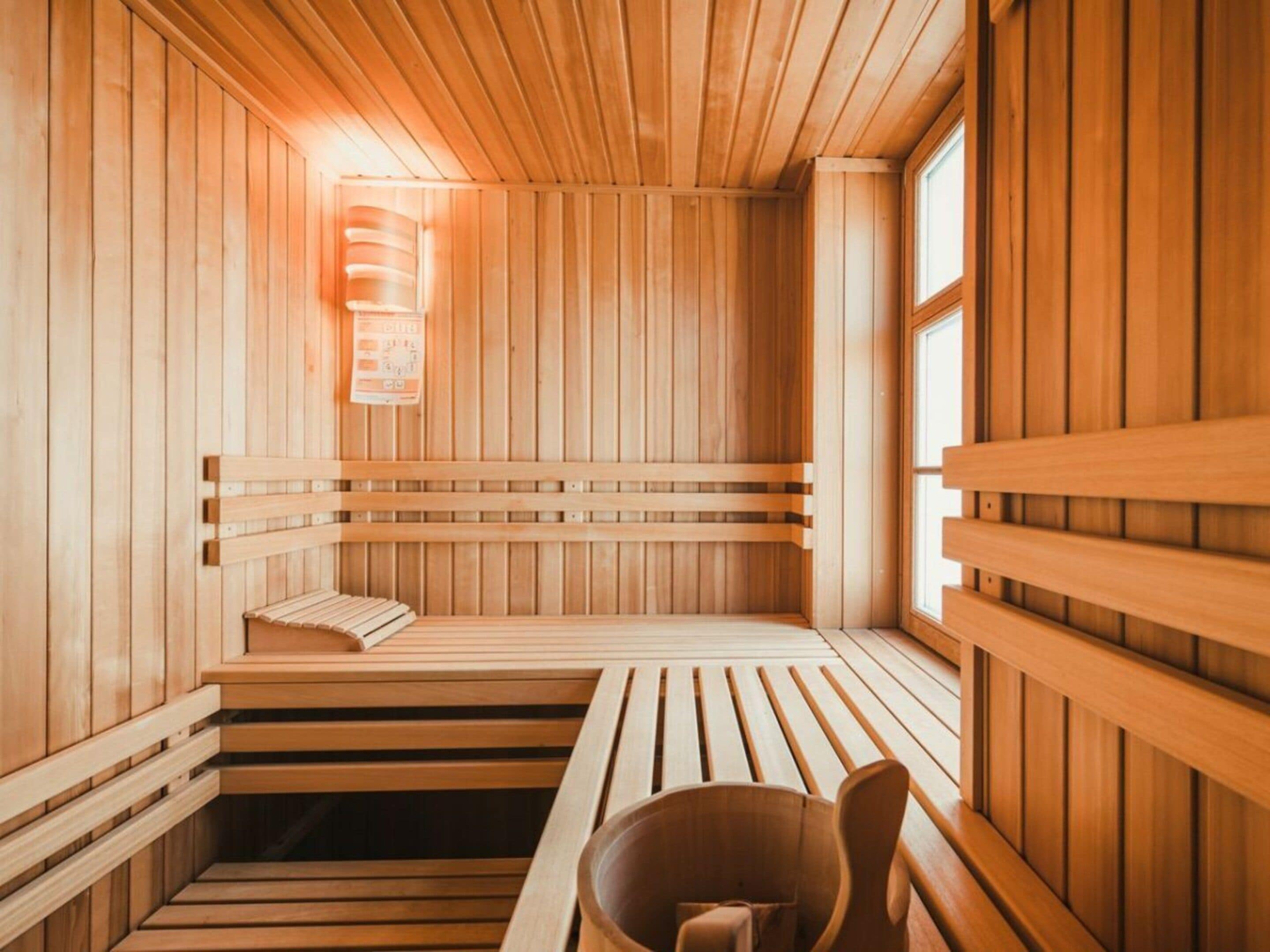 A sauna with wooden benches and a bench seat, a bucket, and a wall-mounted light fixture.