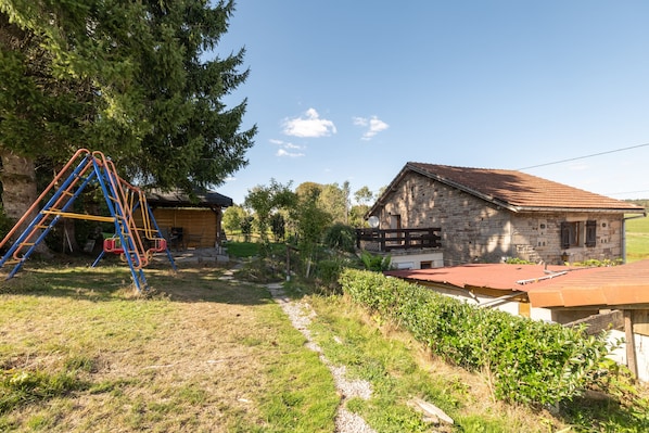 A rustic stone house with a red-tiled roof, a wooden deck, and a swing set in a grassy yard.