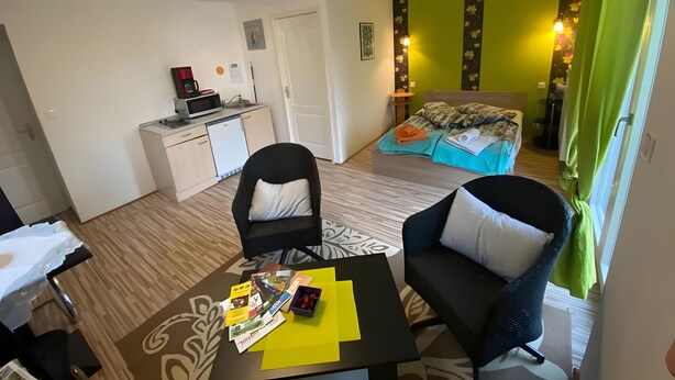 Une chambre d’hôtel compacte comprenant un coin cuisine, un lit avec une literie à motifs floraux, un espace salon avec une table et une vue sur le couloir.