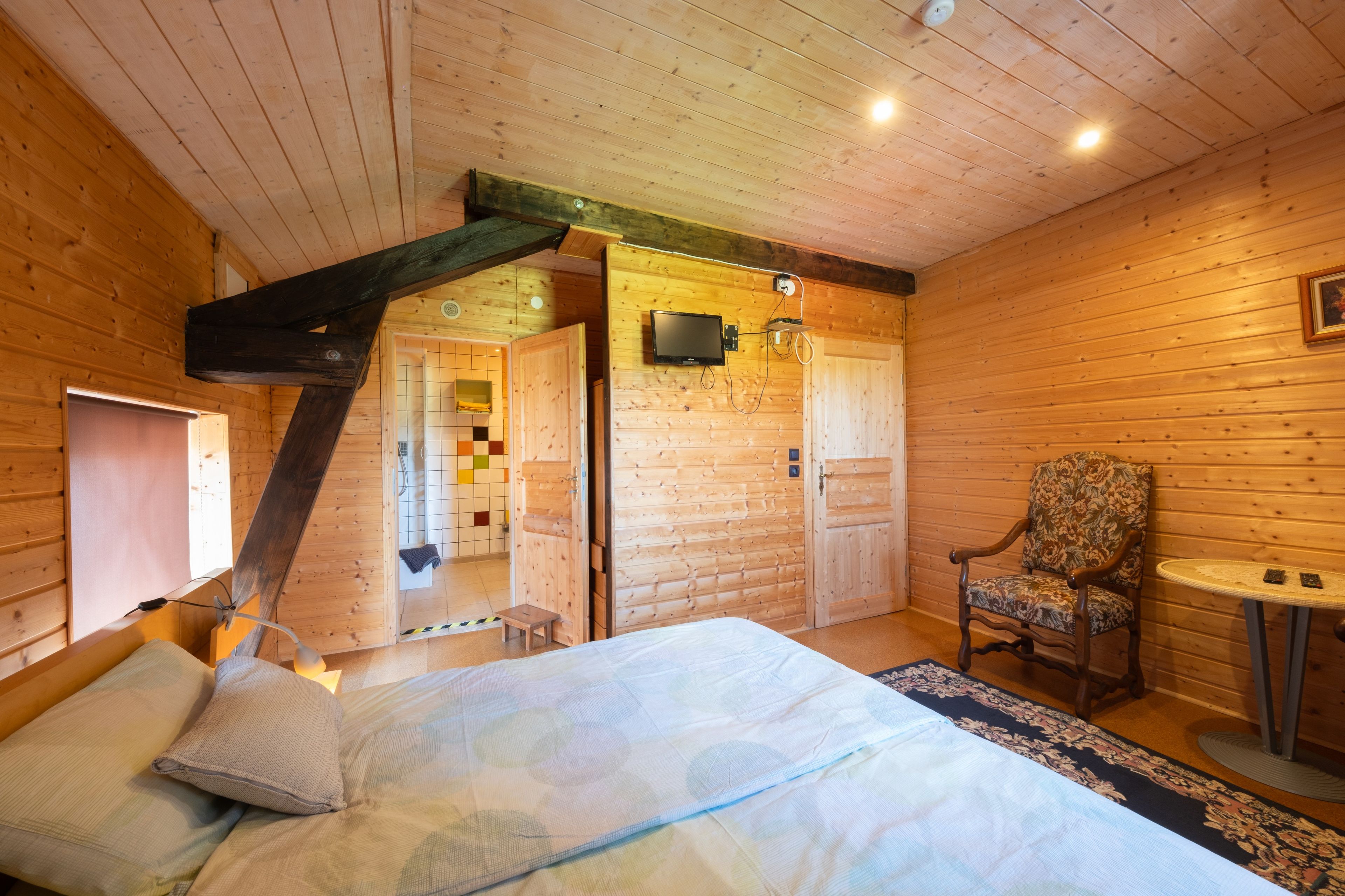 A wooden cabin interior with a bed, a chair, a small table, and a bathroom visible through an open door.