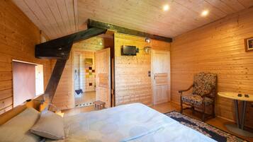 A wooden cabin interior with a bed, a chair, a small table, and a bathroom visible through an open door.