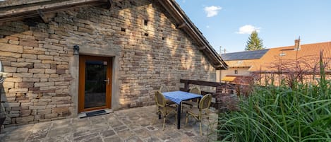 A stone building with a wooden door, a small table with chairs, and a garden area.