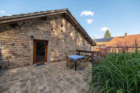 A stone building with a wooden door, a small table with chairs, and a garden area.