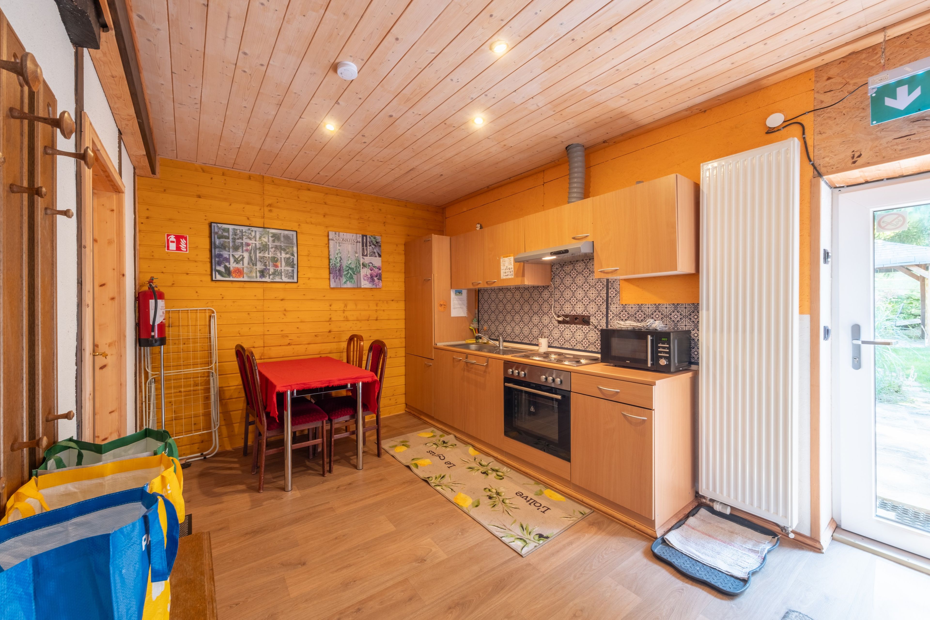 Een kleine keuken met houten kastjes, een magnetron en een rode tafel met stoelen.