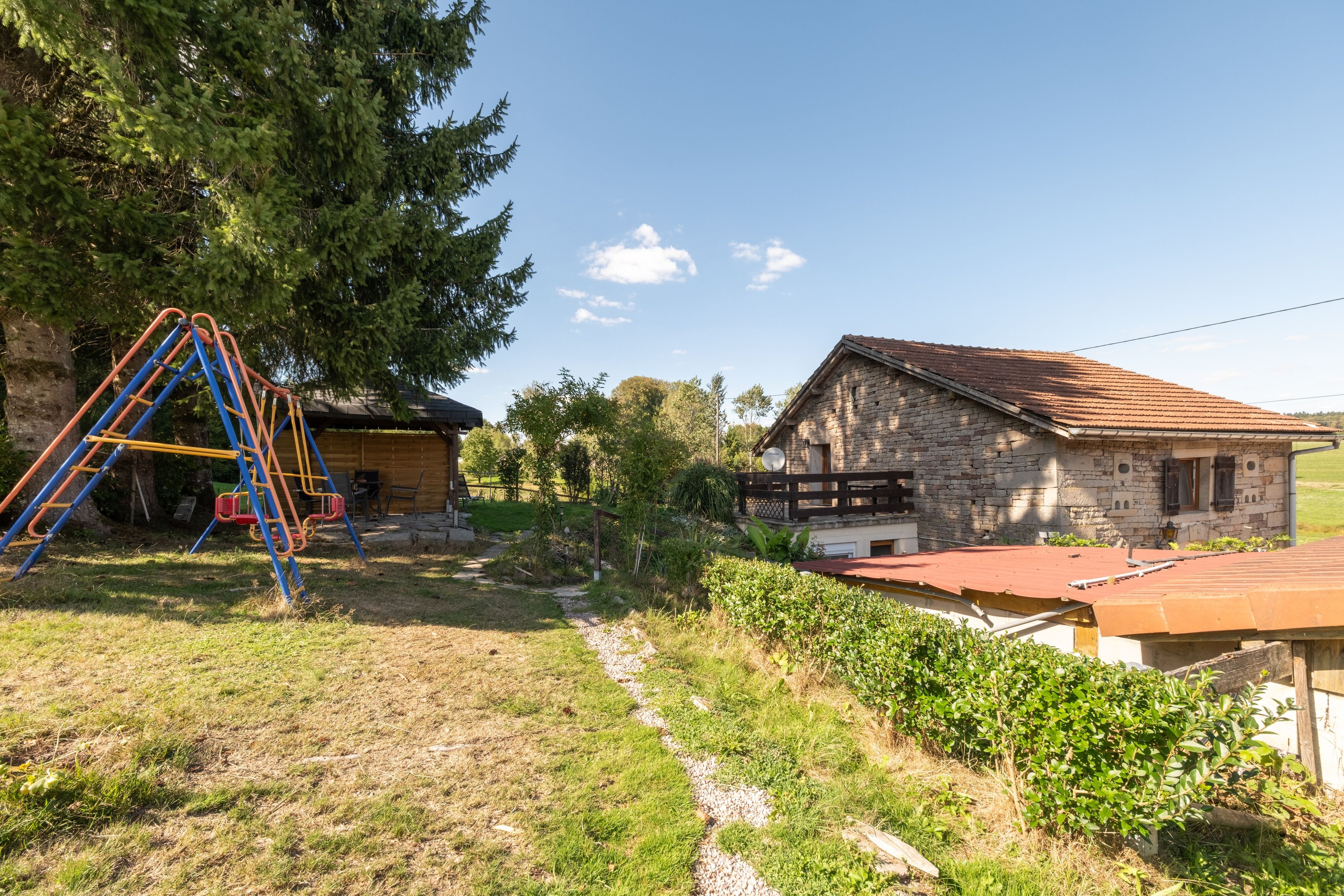 A rustic stone house with a red-tiled roof, a wooden deck, and a swing set in a grassy yard.