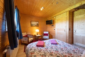 Een houten kamer met een bed, een stoel, een tafel met een lamp, een schilderij aan de muur en een aan de wand bevestigde televisie.