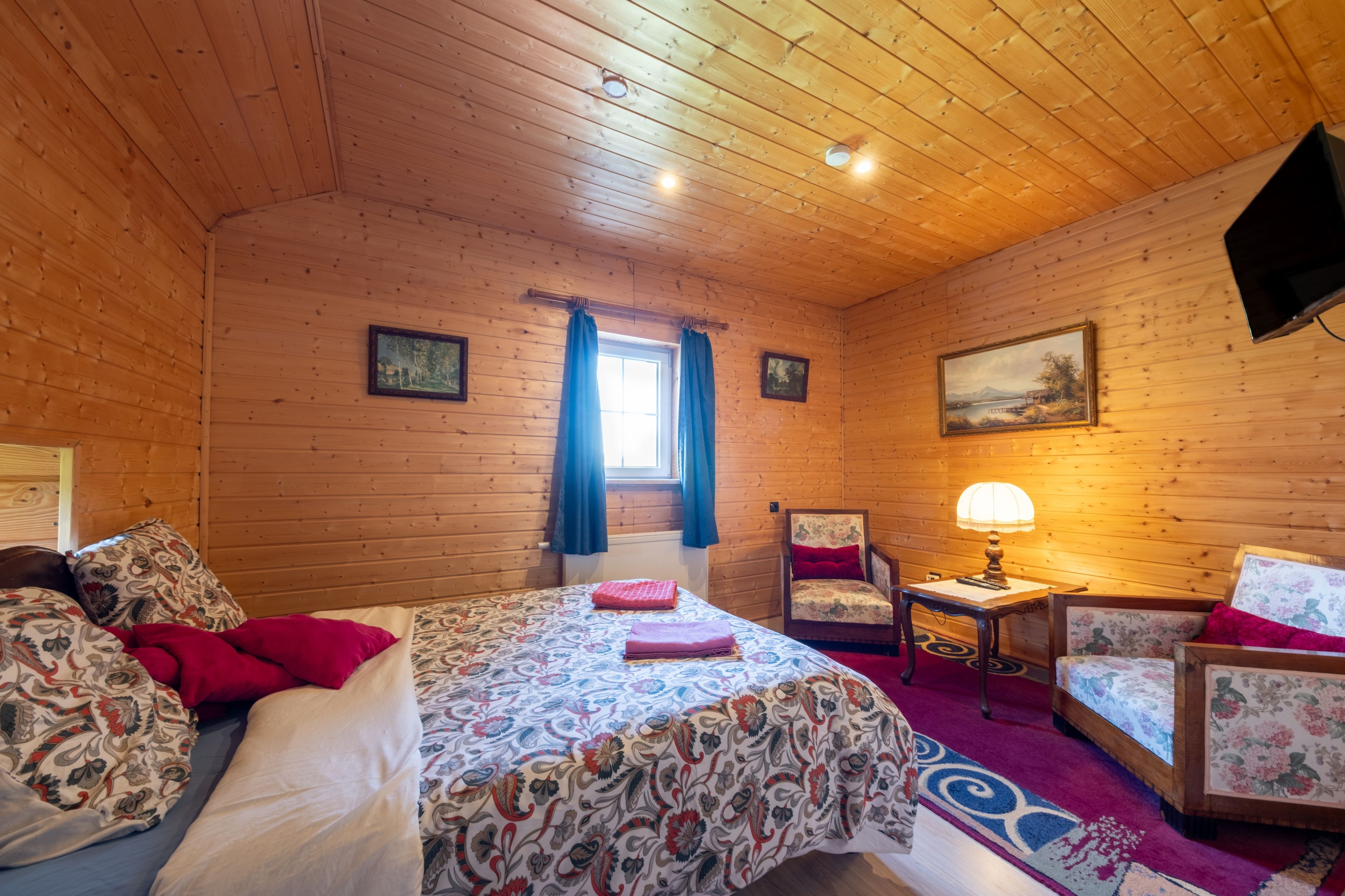 A wooden-paneled room with a bed, a chair, a table with a lamp, and a painting on the wall.