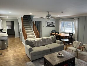 Een woonkamer met een bank, salontafel en een eethoek met stoelen. Een trap leidt naar een hoger niveau.